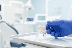 A professional hand in a blue glove holds a white dental crown over a sterile tray in a bright, modern dental clinic. The clean, clinical environment features blurred dental equipment in the background, emphasizing professional oral healthcare and restorative dentistry services.
