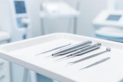 A close-up of professional silver dental instruments and tools arranged on a clean white tray in a bright, sterile dental clinic. The image features dental handpieces and probes against a blurred medical background with soft blue and white tones, highlighting oral healthcare and dentistry.