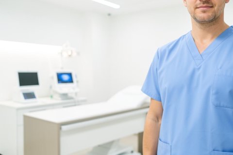 Close-up of a healthcare professional wearing blue medical scrubs in a bright, modern clinical setting. The background shows a clean, well-lit medical examination room equipped with a patient bed and digital diagnostic monitors, creating a professional healthcare environment.