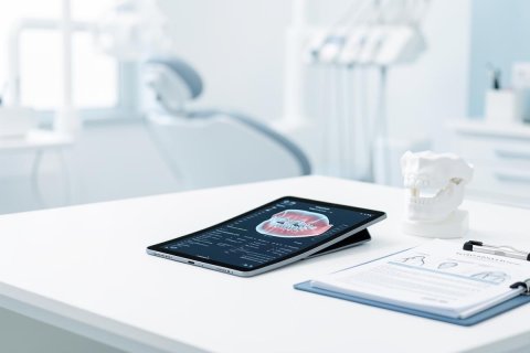 Digital dentistry concept in a bright, modern dental clinic. A tablet on a white desk displays a high-tech 3D scan of teeth and gums, accompanied by an anatomical jaw model and a dental chart clipboard. The sterile, professional environment features soft blue and white tones with blurred dental equipment in the background, highlighting advanced oral healthcare technology.